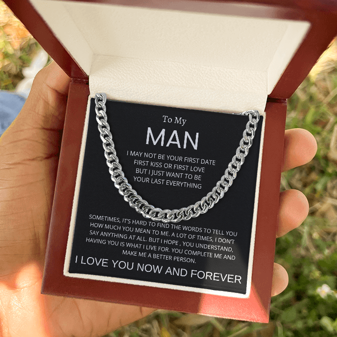 A silver chain necklace presented in an open reddish-brown gift box held in a hand. The necklace is displayed on a black message card addressed 'To My MAN' with a romantic quote, against an outdoor blurred background.