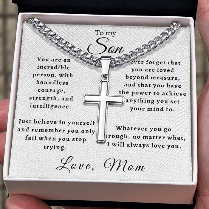 A silver cross necklace on a silver chain presented in an open gift box with a printed message for a son, held in a person's hand.