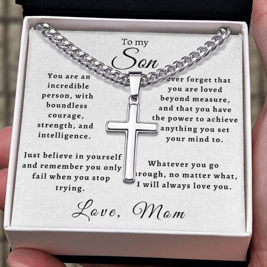 A silver cross necklace on a silver chain presented in an open gift box with a printed message for a son, held in a person's hand.